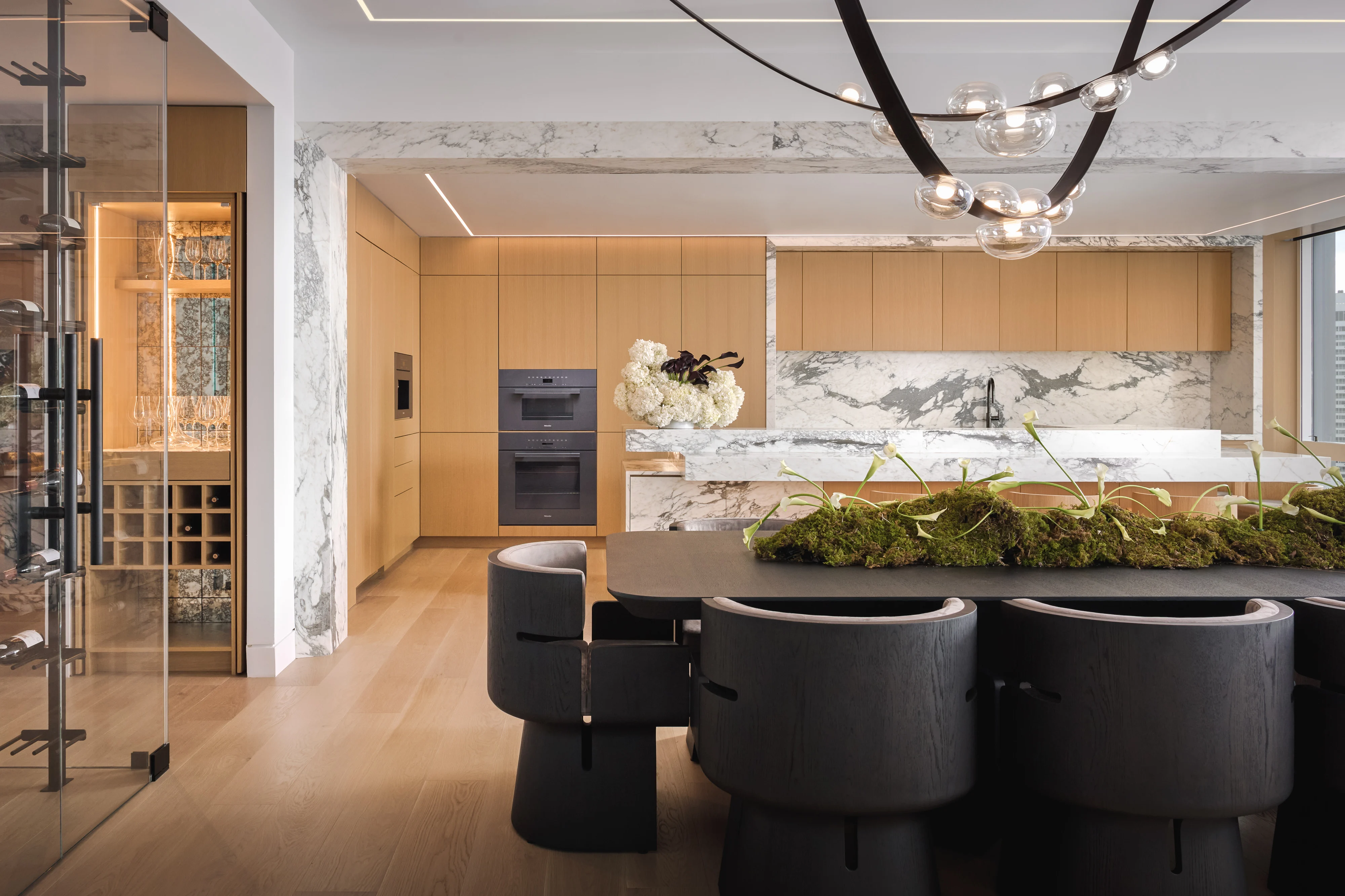 Oak kitchen with a marble island and backsplash beyond a dining table set under a looping chandelier