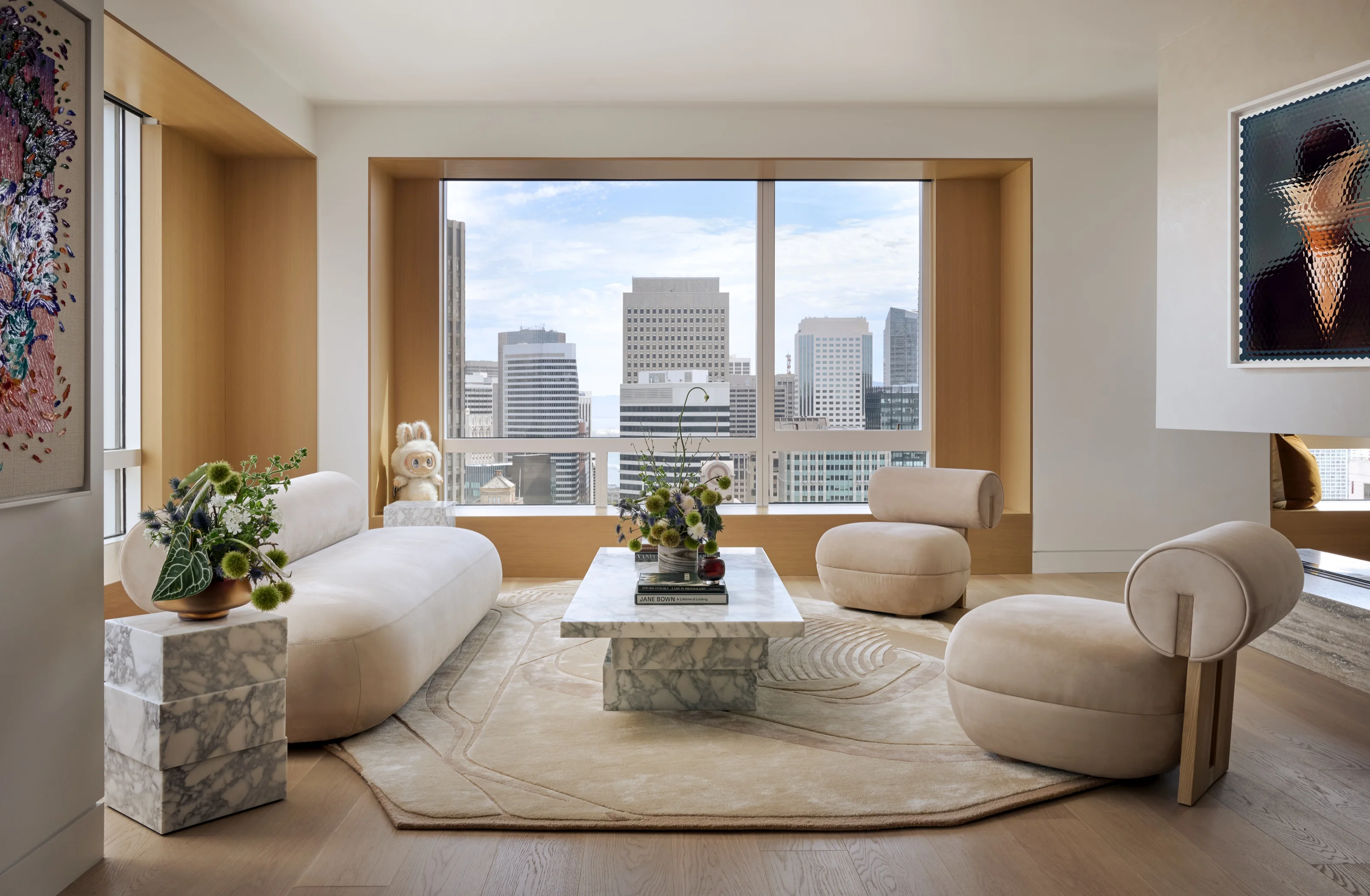 Living room with marble block tables and cream boucle seating before a wall of windows over the city