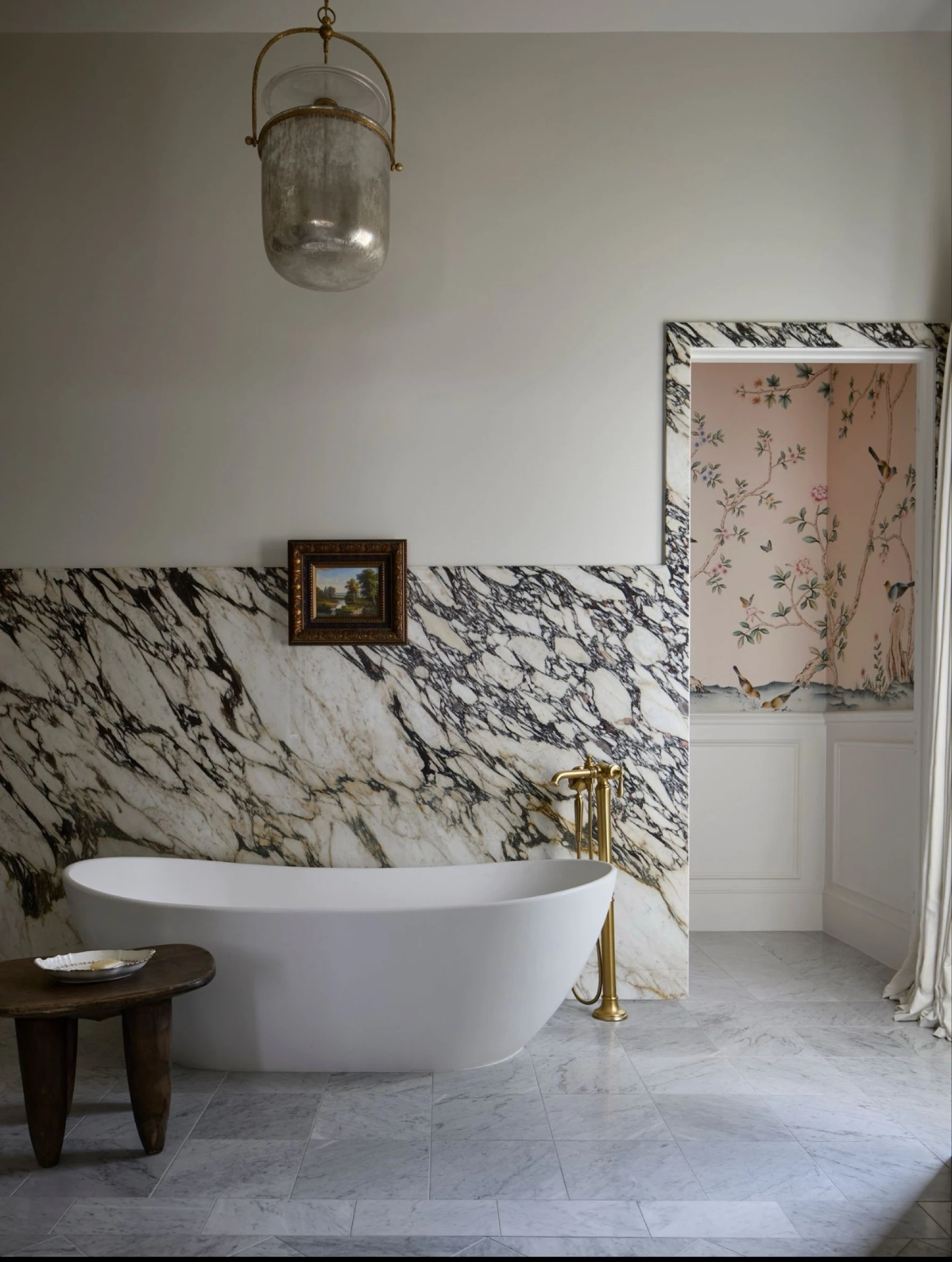 Freestanding tub against a wall of dramatically veined marble, chinoiserie wallpaper in the alcove beyond