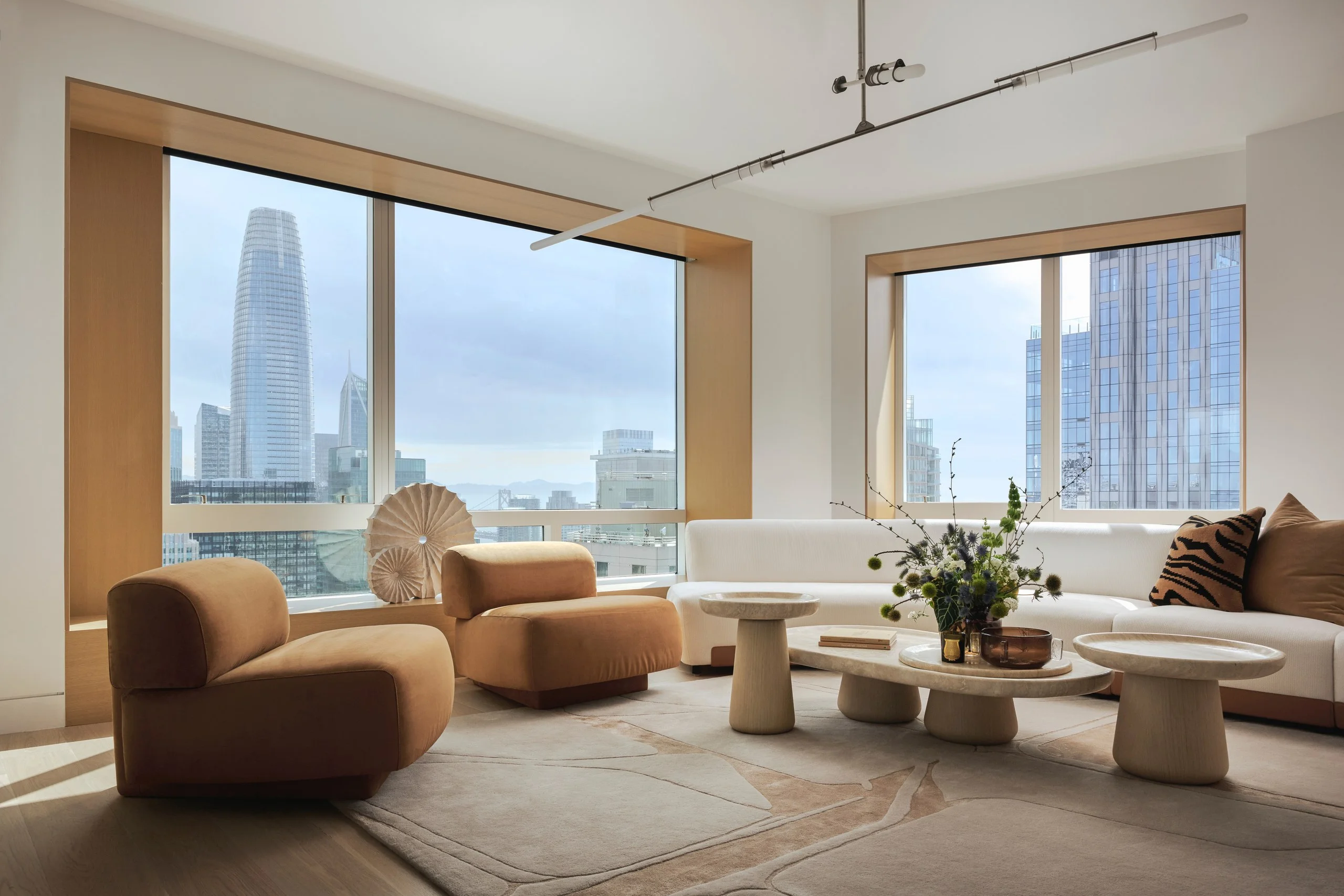 Living room high above a city, with corner windows, curved tan armchairs and a cream sectional around low stone tables